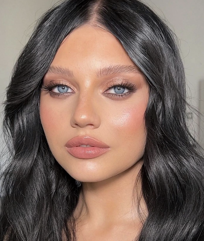 A woman with a sultry smokey eye makeup look, highlighted by shades of warm brown and soft black, giving her blue-grey eyes a dramatic effect. Her skin glows with a natural-looking, dewy finish, accentuated with a touch of rose blush on the cheeks. Her full lips are painted in a matte nude tone, complementing the overall muted color scheme. Her jet-black hair is styled in loose waves, framing her face beautifully and completing the captivating look.